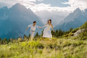 An adventurous couple holds hands and laughs while running down a sunny alpine meadow, with the dramatic mountains of the French Alps behind them, in a photo by their Chamonix couple photographer.