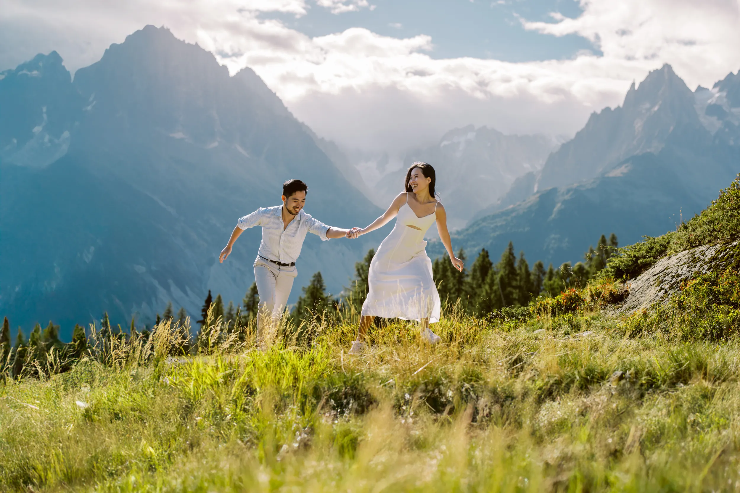 An adventurous couple holds hands and laughs while running down a sunny alpine meadow, with the dramatic mountains of the French Alps behind them, in a photo by their Chamonix couple photographer.