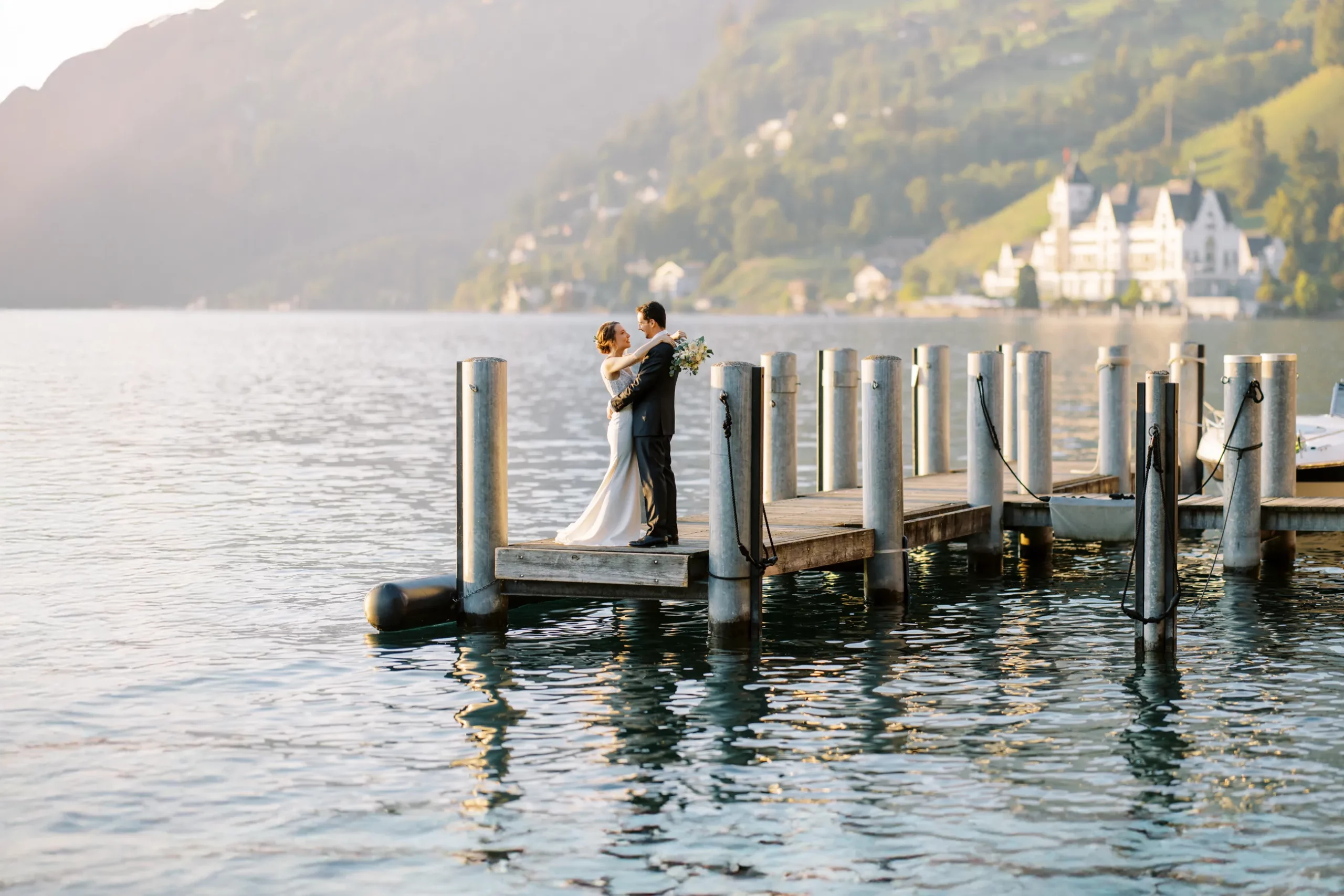 A bride and groom embrace on a wooden pier on a lake in Switzerland, with majestic mountains in the background, a moment captured by their wedding photographer in Switzerland.