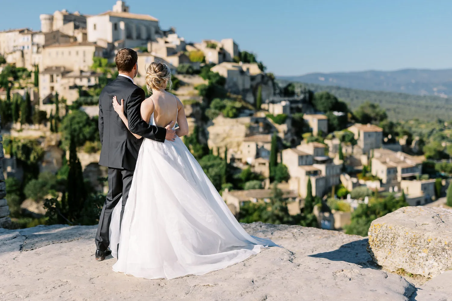 A bride and groom stand on a rocky overlook with their backs to the camera, looking at the historic village of Gordes, a scene captured by their wedding photographer in Provence.