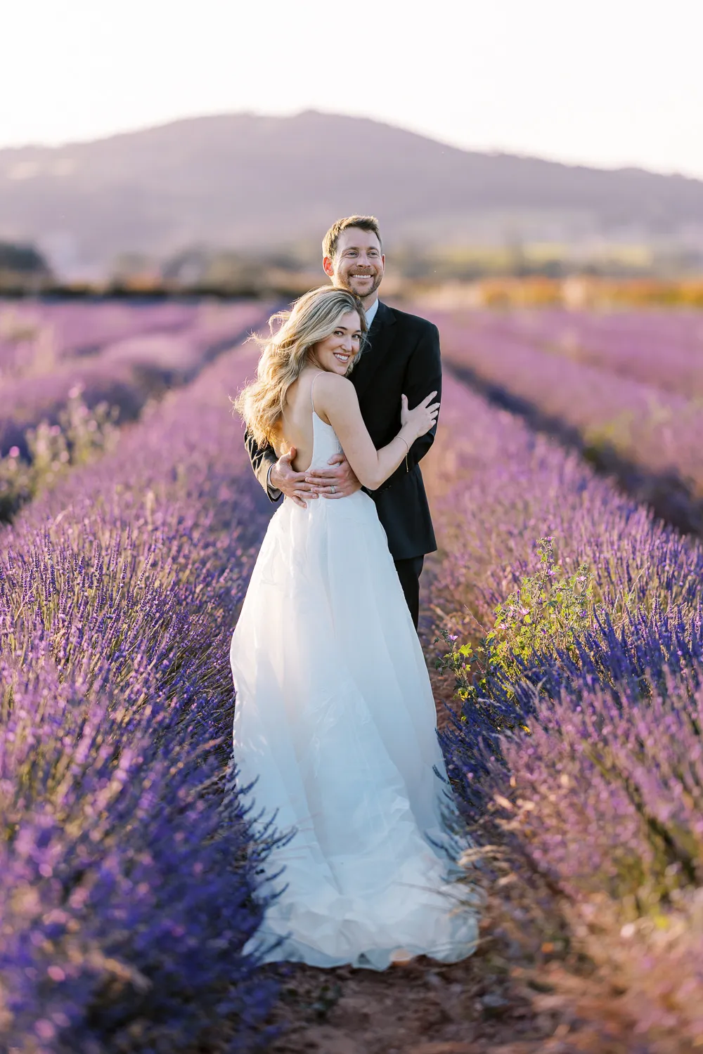 A joyful bride, embraced by her groom from behind, smiles at the camera while standing in the middle of a stunning lavender field at sunset in Provence.