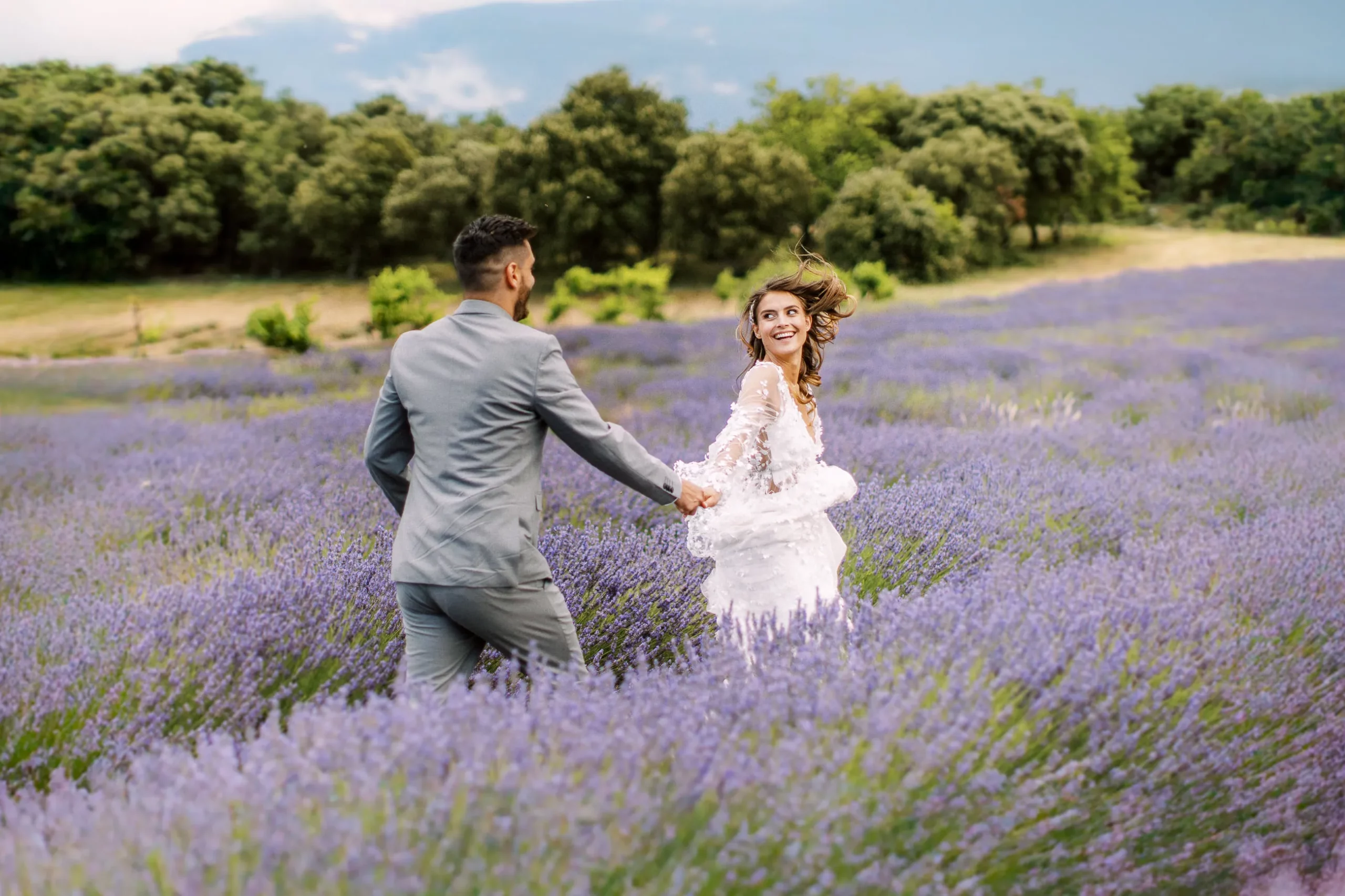Bride looking back and smiling while groom leads her through a vibrant lavender field in Provence - captured by wedding photographer Provence.