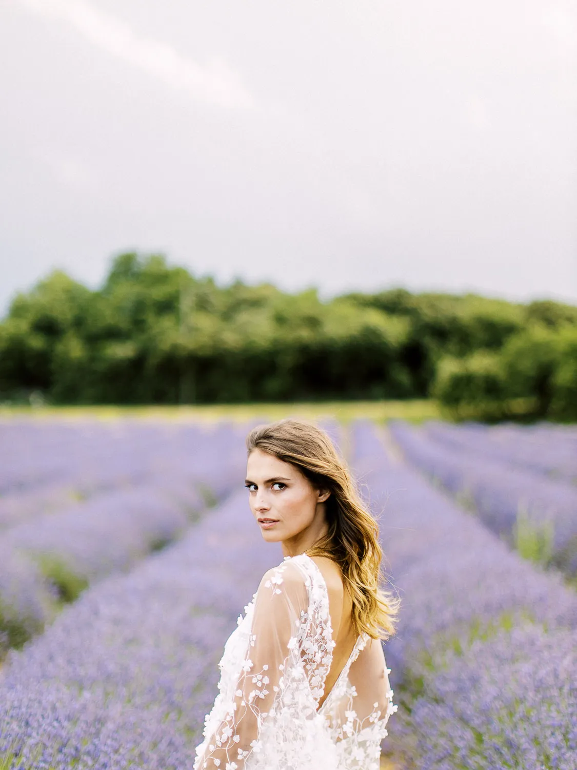 Beautiful bride in a lace dress looking over her shoulder, standing in a blooming Provence lavender field - portrait by wedding photographer Provence.