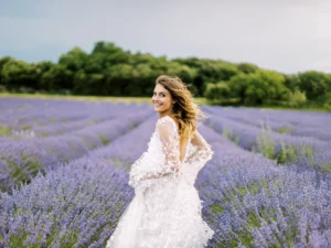 A joyful bride in a lace wedding dress smiles over her shoulder while standing in a blooming lavender field, in a photo by a wedding photographer in Provence.