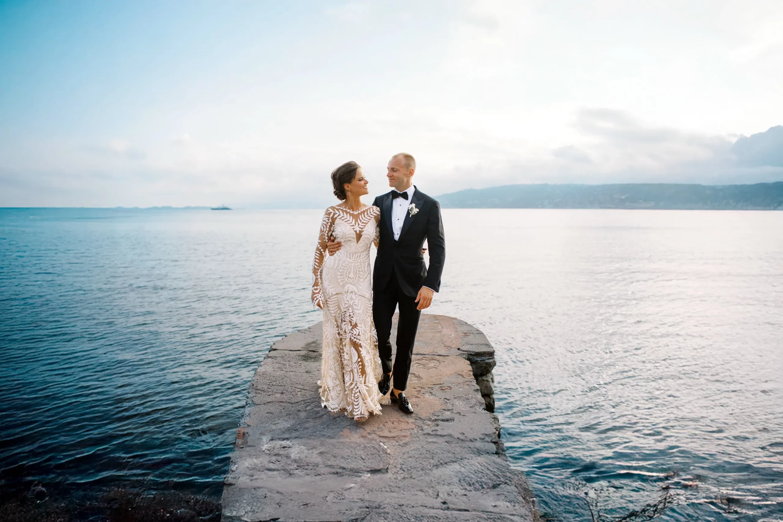 An elegant bride and groom walk on a stone pier overlooking the Mediterranean Sea, a romantic moment captured by their wedding photographer in Malta.