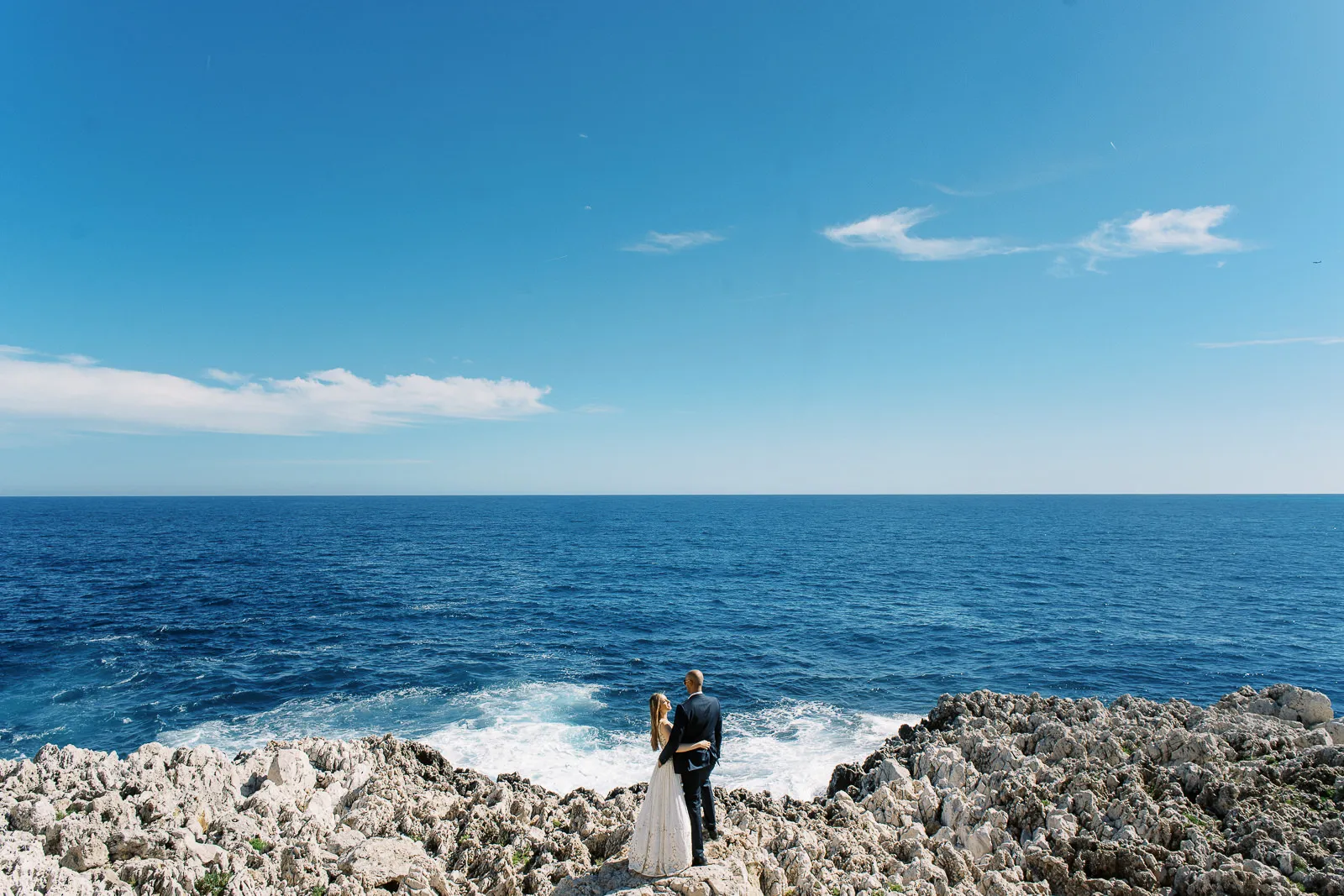 Bride and groom standing on a dramatic rocky coastline overlooking the blue Mediterranean Sea - epic shot by wedding photographer French Riviera.
