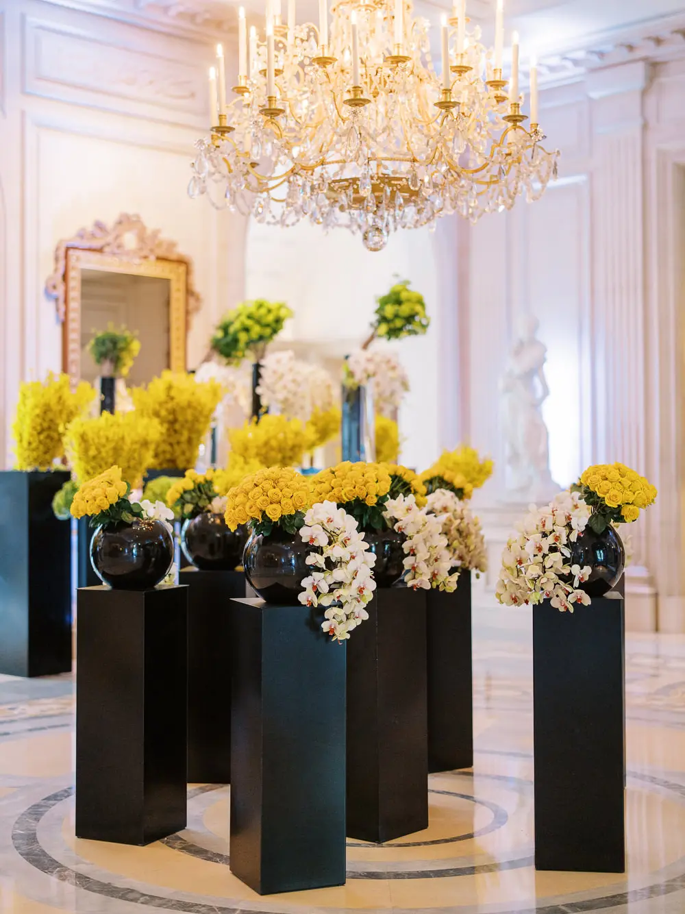 Modern chic wedding decor with yellow flowers and white orchids in black vases on pedestals under a chandelier - Geneva wedding photographer shot.