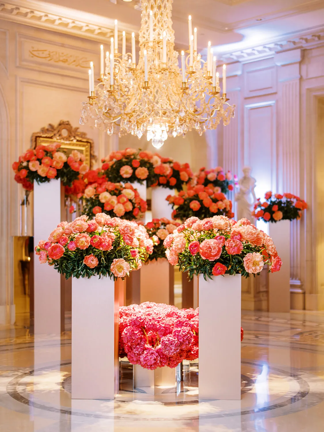 Opulent wedding reception decor with large pink and coral peony arrangements on white pedestals under a grand chandelier - photographed by wedding photographer Geneva.