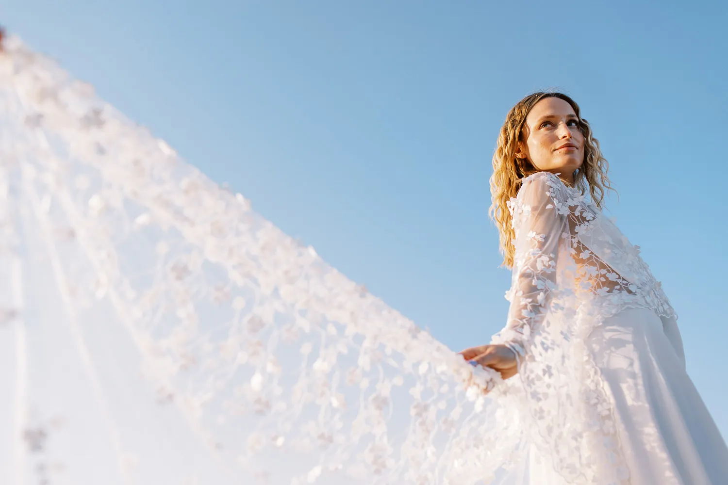 Ethereal bride holding a long lace veil against a clear blue Maltese sky - photo by wedding photographer Malta.