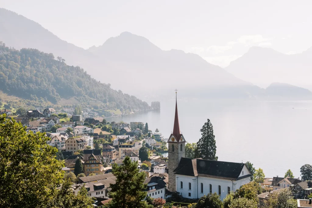 A church on a hill overlooks a lake and mountains, with a couple posing in front of Lake Geneva with a wedding photographer Switzerland.