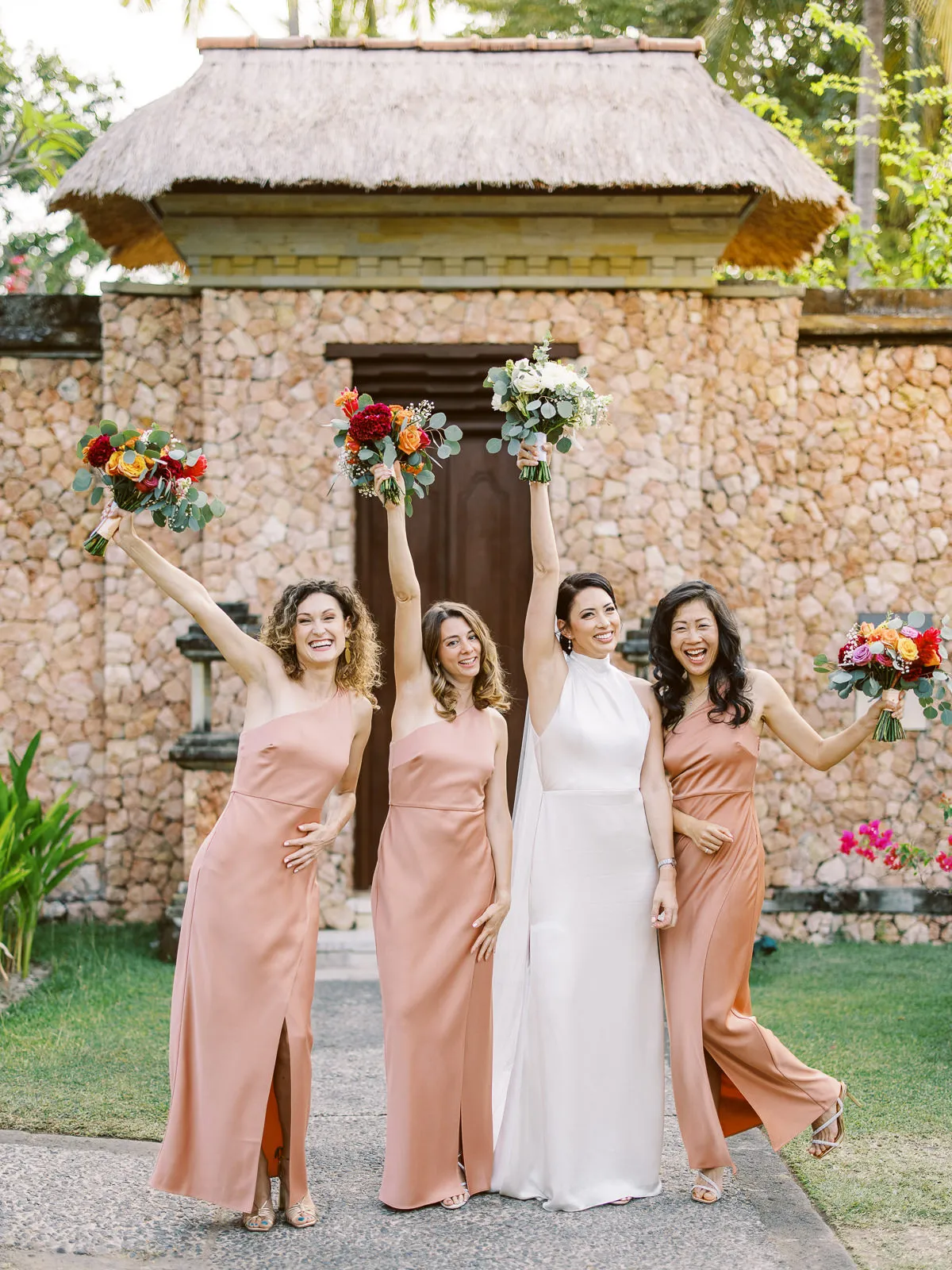 Happy bride and bridesmaids in terracotta dresses cheering with bouquets outside a Lombok villa - photo by wedding photographer Lombok.