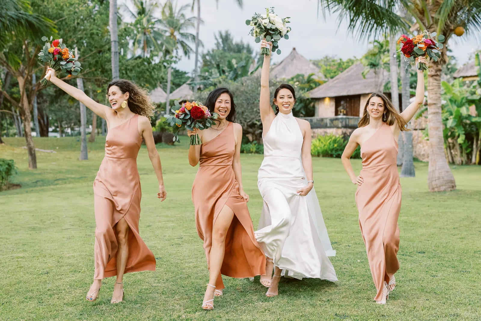 Happy bride and bridesmaids in terracotta dresses running and cheering on a lawn in Lombok - photo by wedding photographer Lombok.