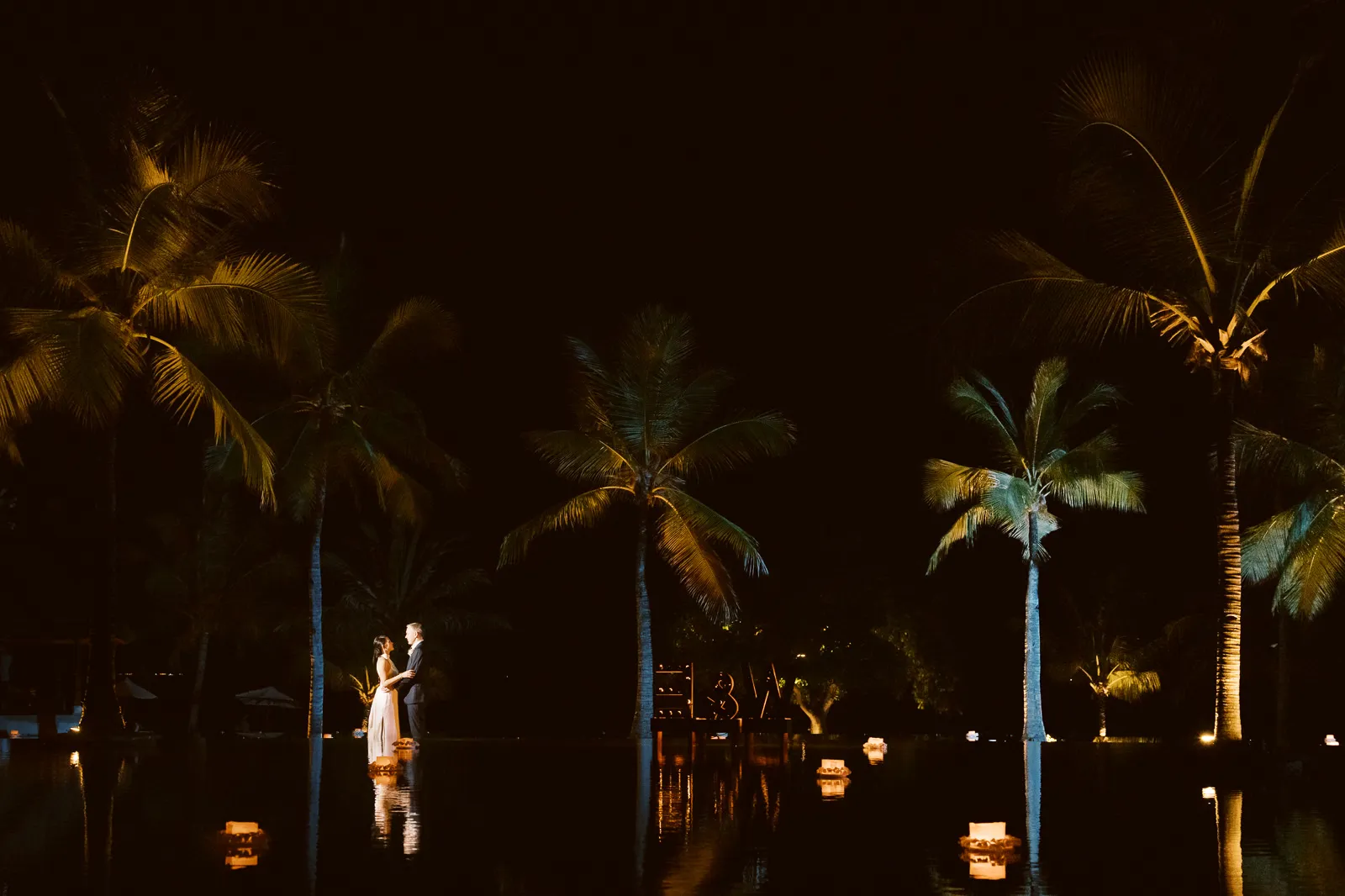 Bride and groom standing by illuminated pool surrounded by palm trees at night in Lombok - photo by wedding photographer Lombok.