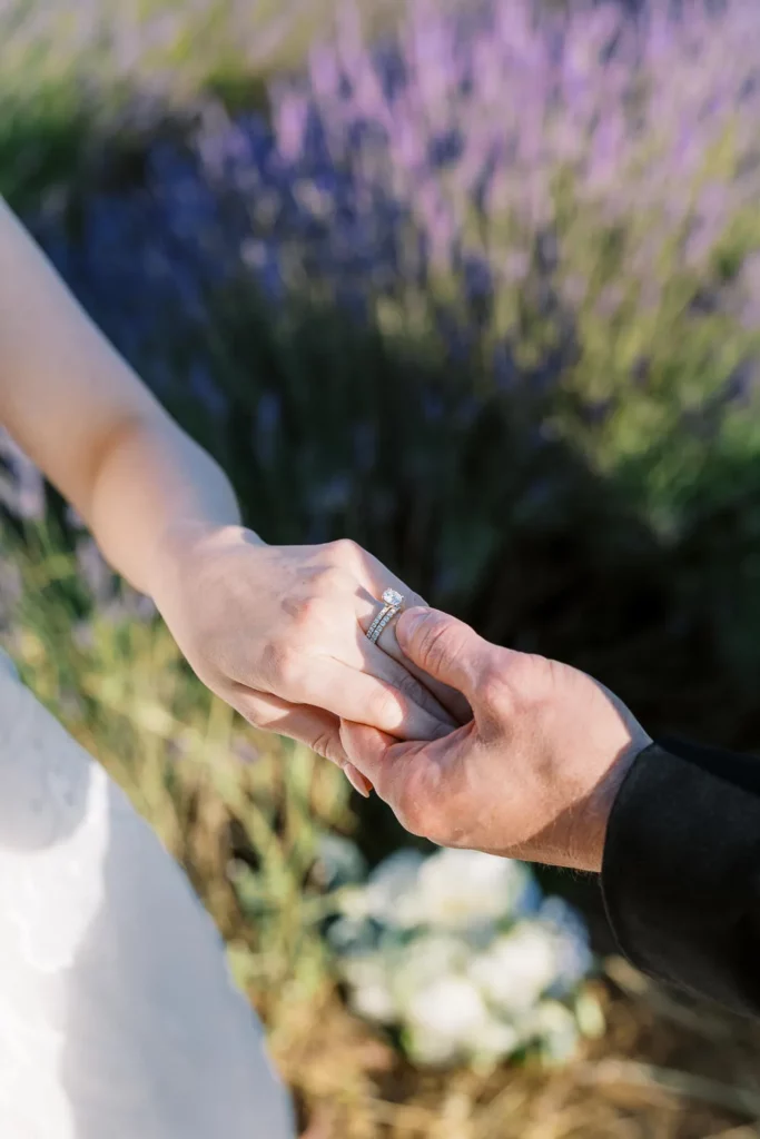 A close-up shot focusing on a bride's diamond engagement ring as she holds hands with her fiancé in a lavender field.