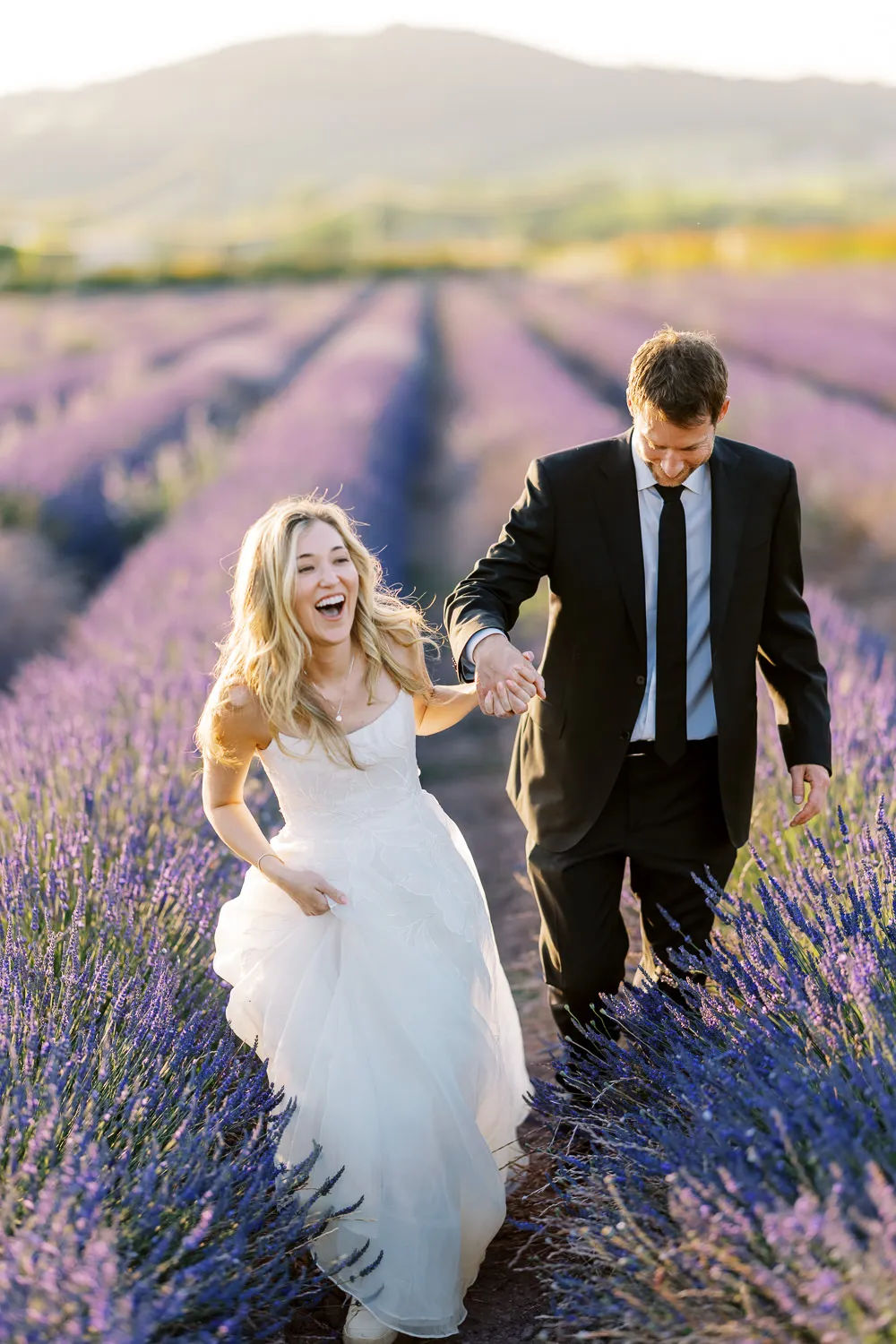 A joyful bride laughs while holding her groom's hand and walking through a purple lavender field, captured by their wedding photographer in Provence. Lavender couple photo.