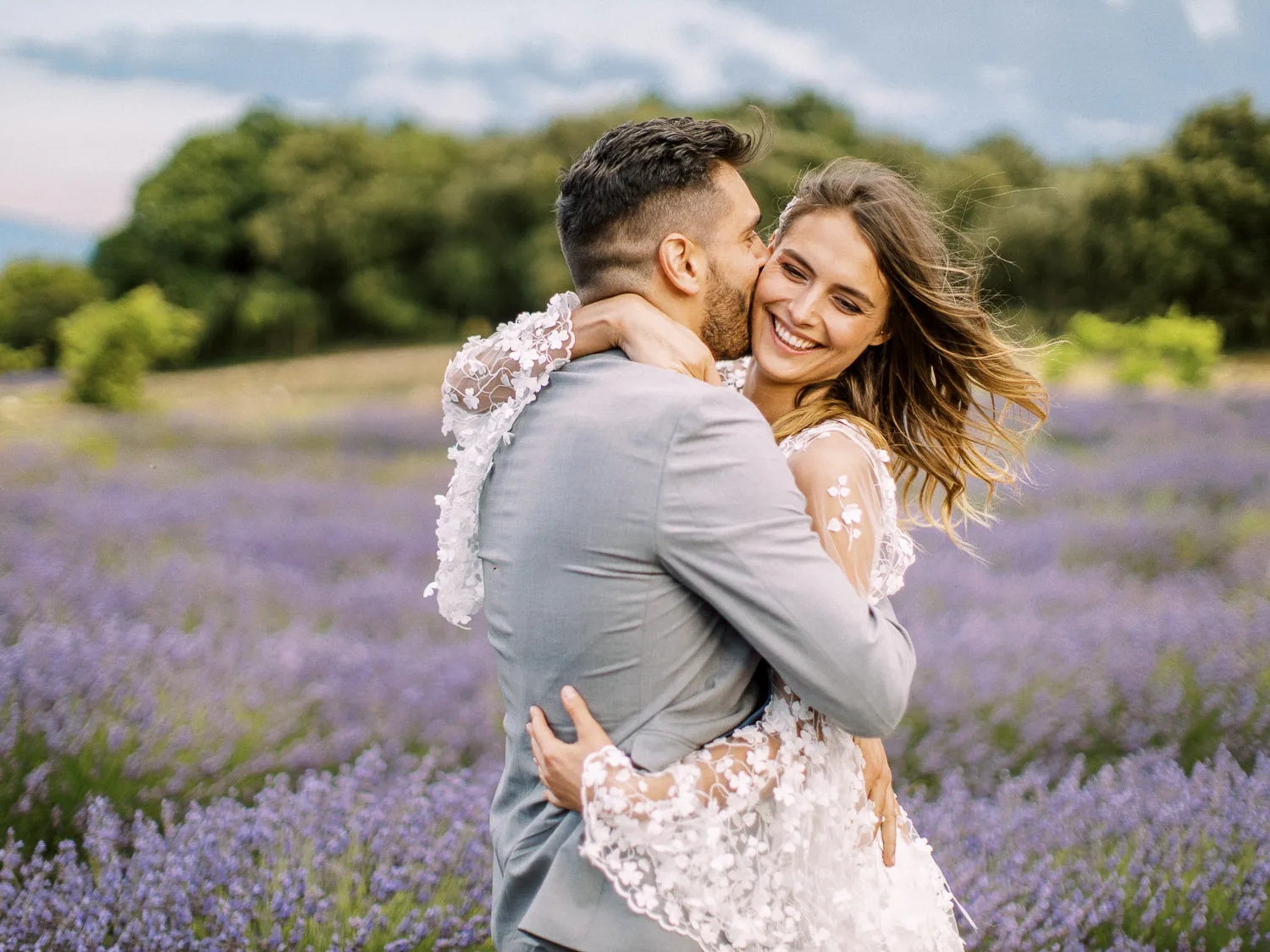 A groom in a light grey suit joyfully kisses his smiling bride on the cheek as they embrace in a vibrant lavender field in Provence. Lavender couple photo.
