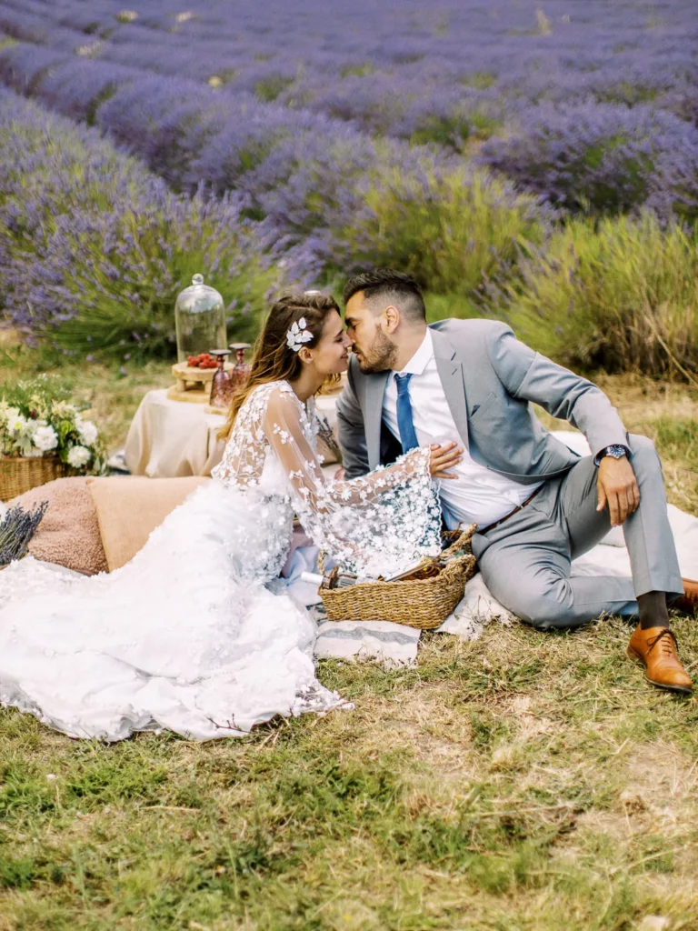 A bride and groom in wedding attire lean in to kiss while enjoying a romantic picnic on a blanket in a lavender field. Lavender couple photo.