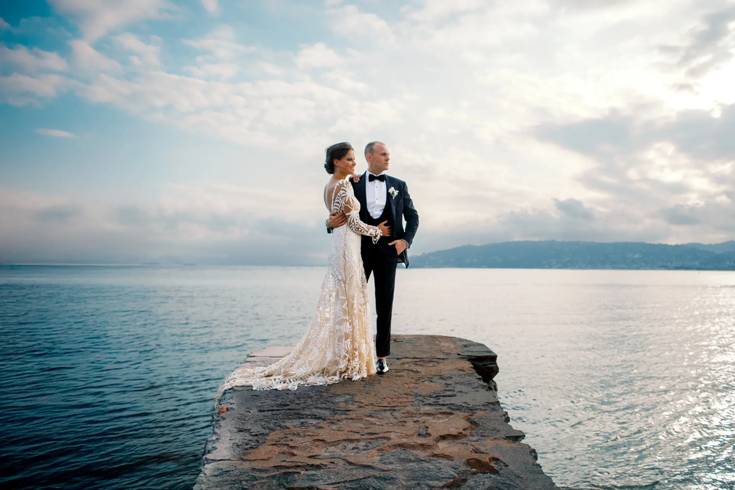 A bride and groom stand together on a stone pier against a dramatic sky, in an epic photo by a wedding photographer in Saint Tropez.