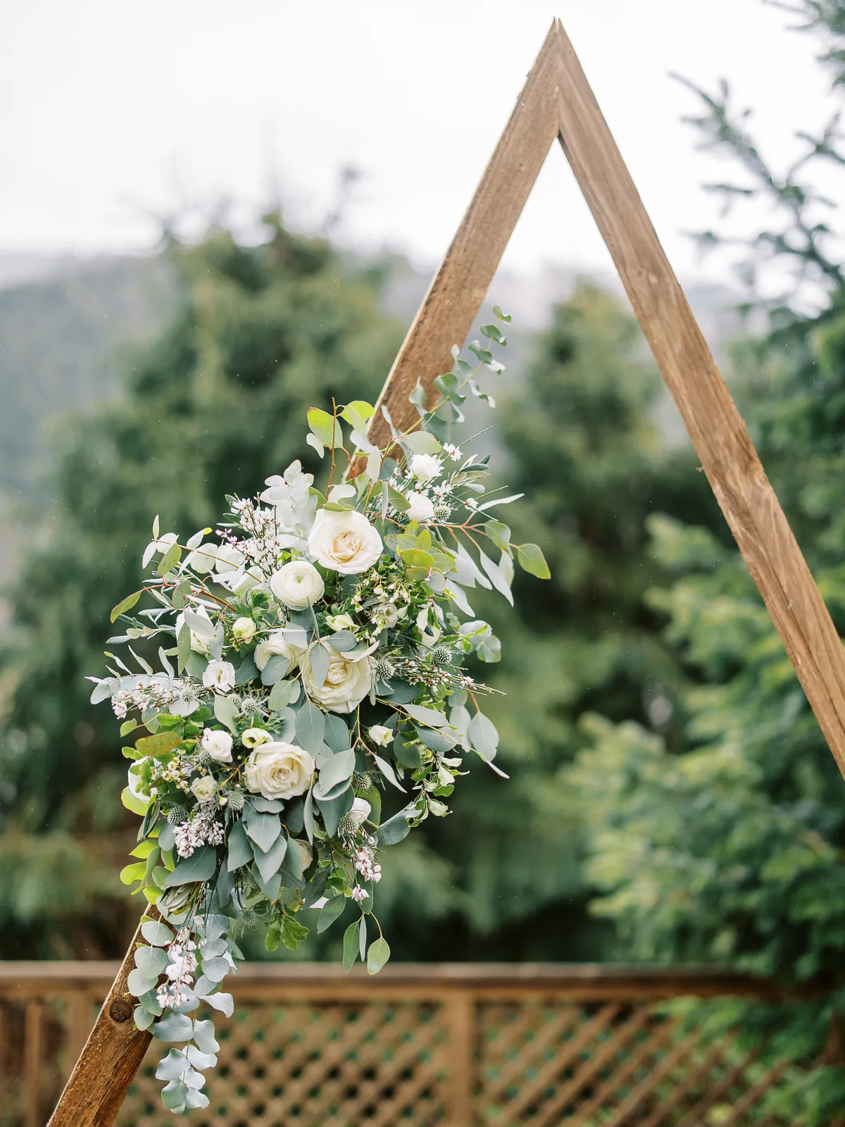 A close-up of a modern wooden triangle wedding arch decorated with white roses and eucalyptus for a French Alps wedding.
