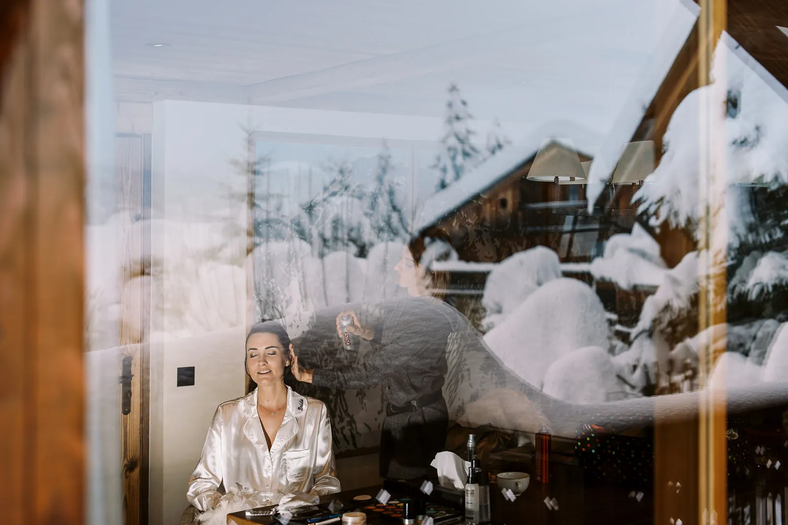 An artistic photo by a Megeve wedding photographer showing a bride getting her makeup done, with a snowy French Alps chalet scene reflected in the window.