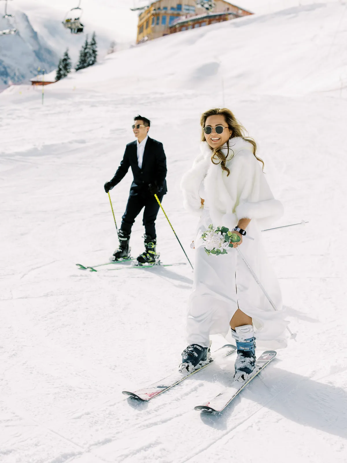 A fun bride and groom in full wedding attire skiing down a slope in Megeve, a unique shot by their adventurous French Alps wedding photographer.
