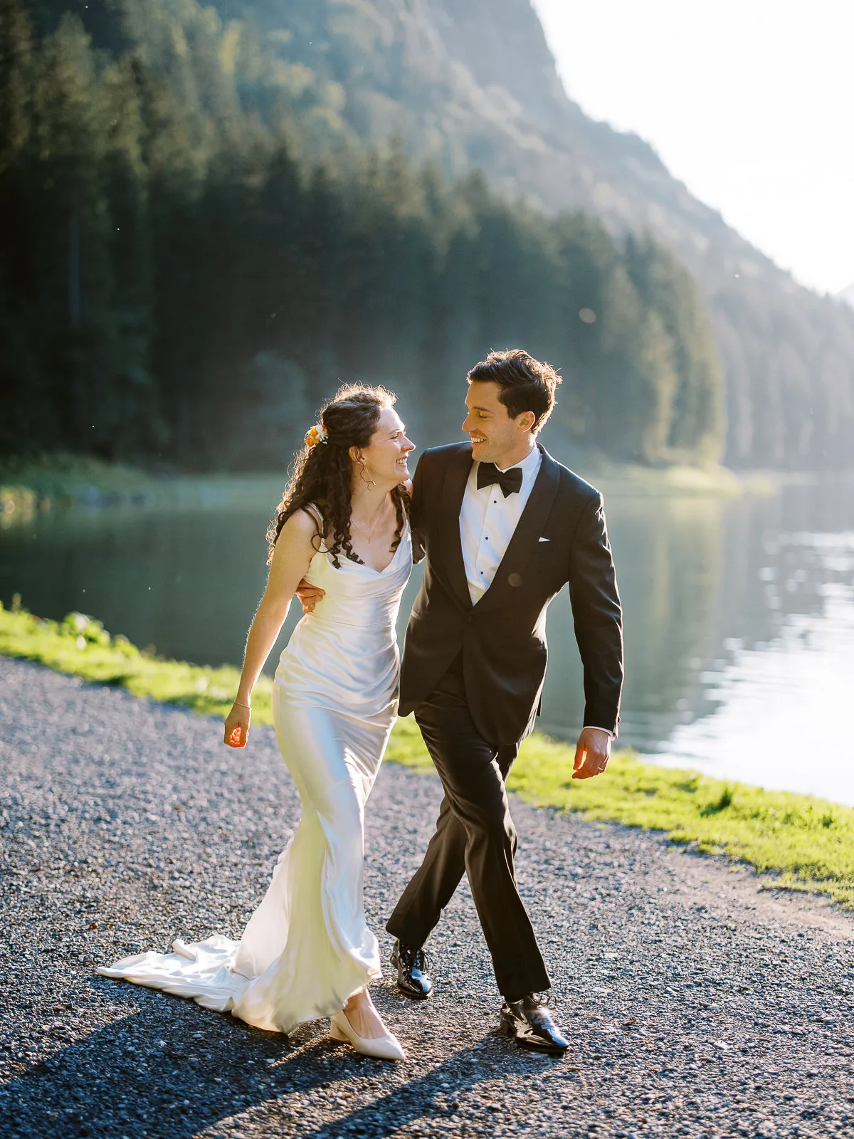 A joyful bride and groom walk along a lakeside path during golden hour, a candid moment captured by their wedding photographer in the French Alps near Megeve.