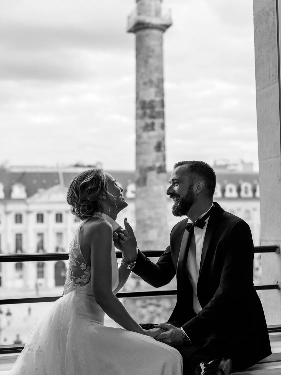 Black and white photo of bride and groom laughing on a Parisian balcony, Place Vendôme column visible - by Wedding photographer Ritz Paris. Photographe mariage Ritz Paris