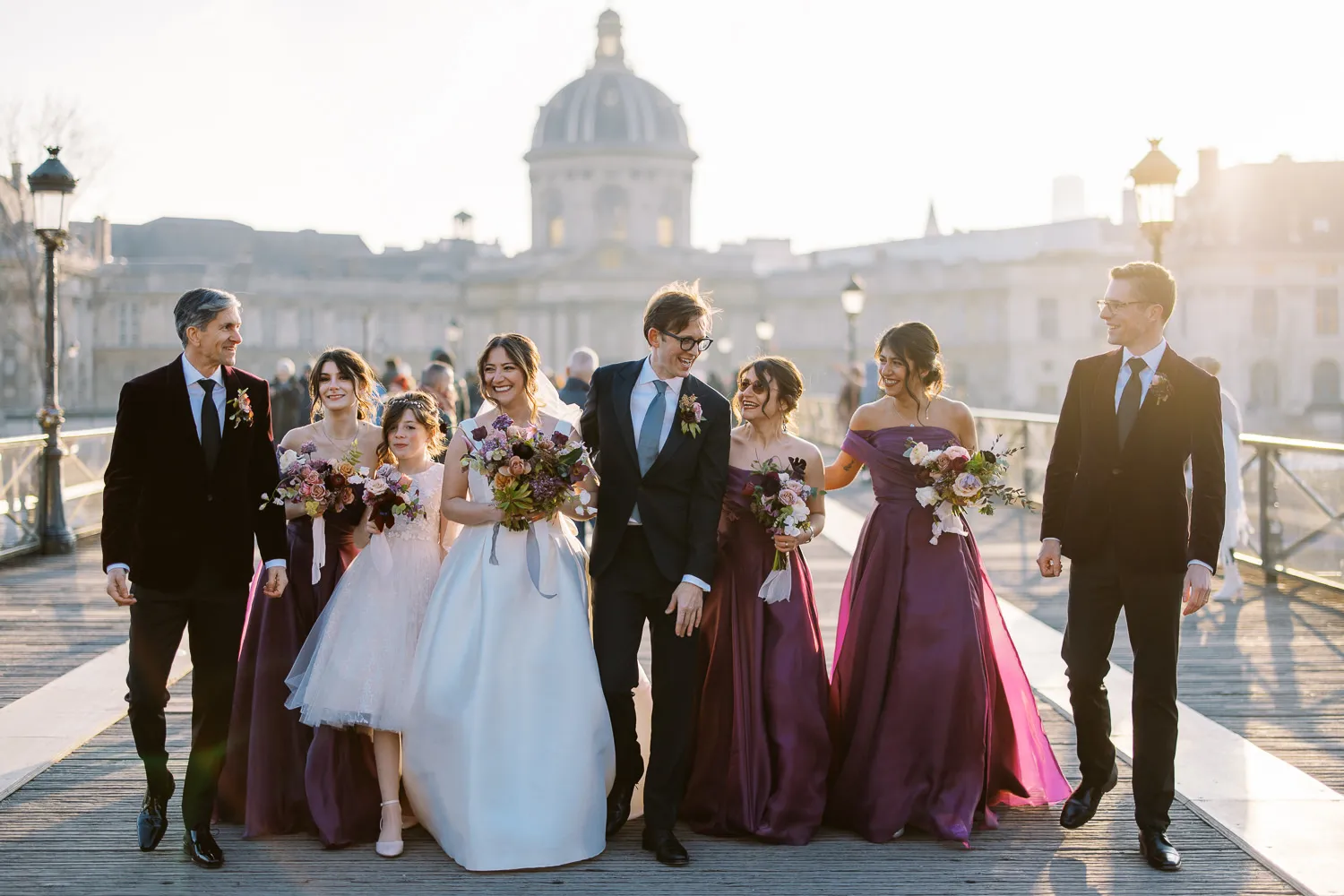 Joyful wedding party walking across Pont des Arts in Paris with Institut de France in background - photo by Wedding photographer Ritz Paris.