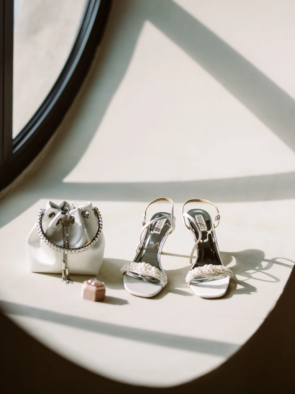 A bride's elegant accessories, including pearl-strapped high heels and a satin pouch bag, are artistically arranged in a beam of sunlight.