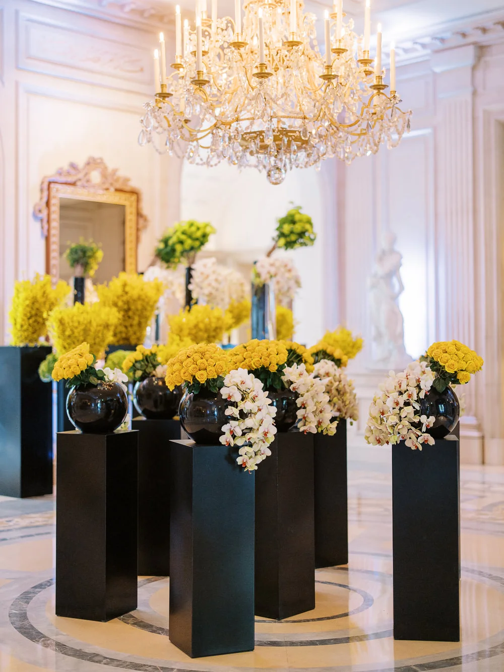A magnificent floral installation with yellow and white flowers in black vases on pedestals under a grand chandelier in a luxury hotel George V Paris.