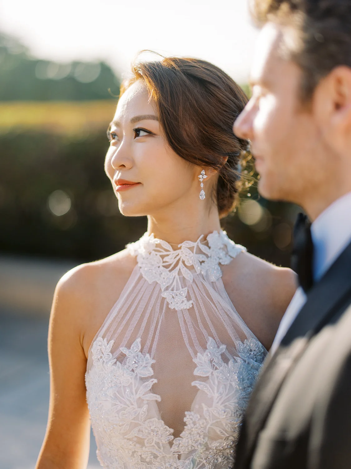 A beautiful bride with an elegant updo and intricate high-neck lace gown looks off-camera during golden hour.