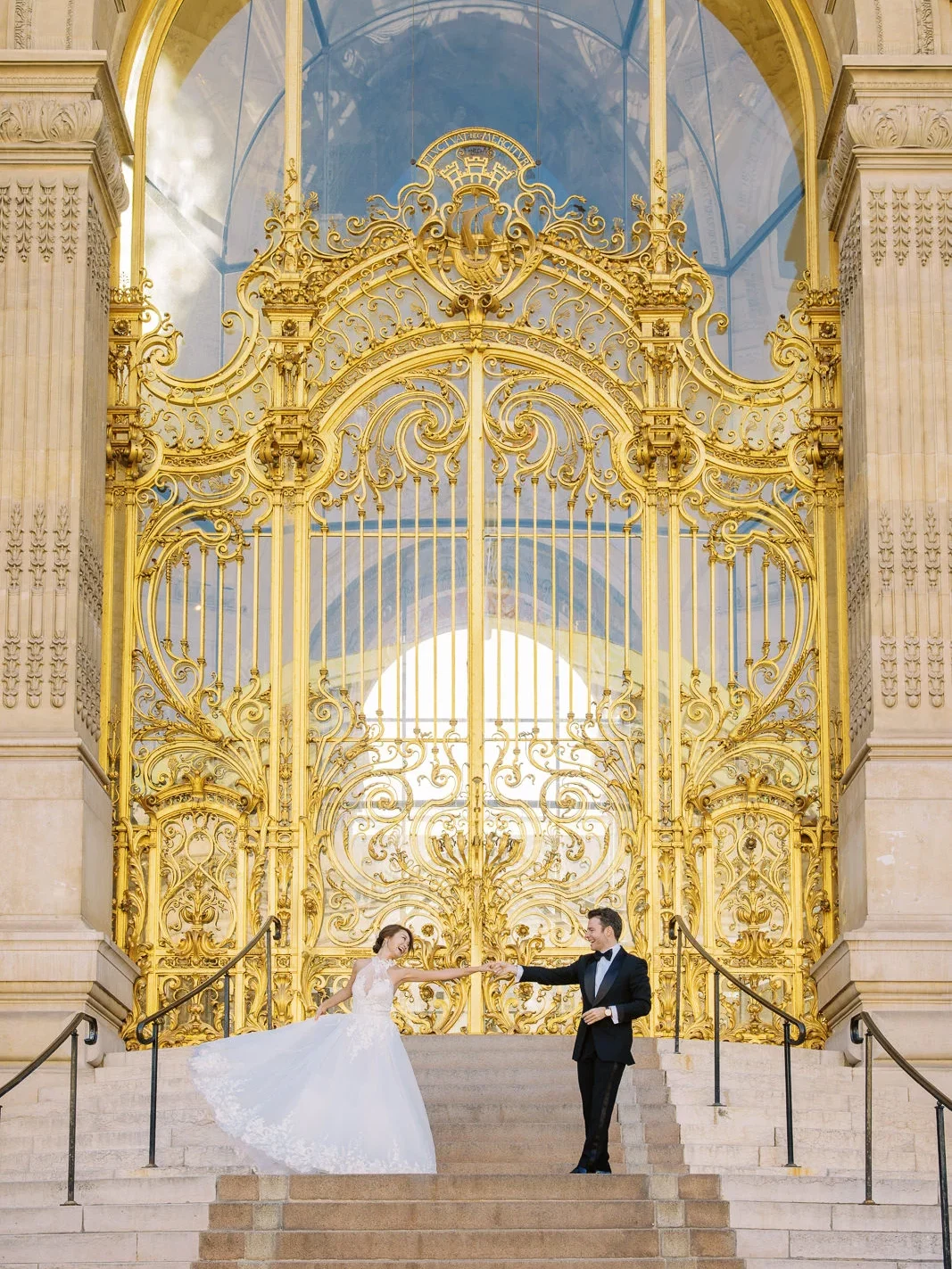 A bride and groom hold hands and dance down the steps of the Petit Palais, with its grand golden doors behind them. How Much Does a Photographer in Paris Cost?