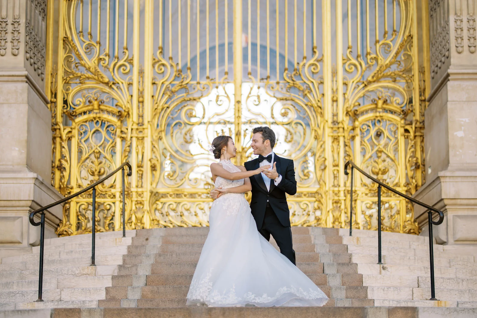 A bride and groom share a joyful moment, about to dance on the steps in front of the magnificent golden gates of the Petit Palais in Paris. How Much Does a Photographer in Paris Cost?