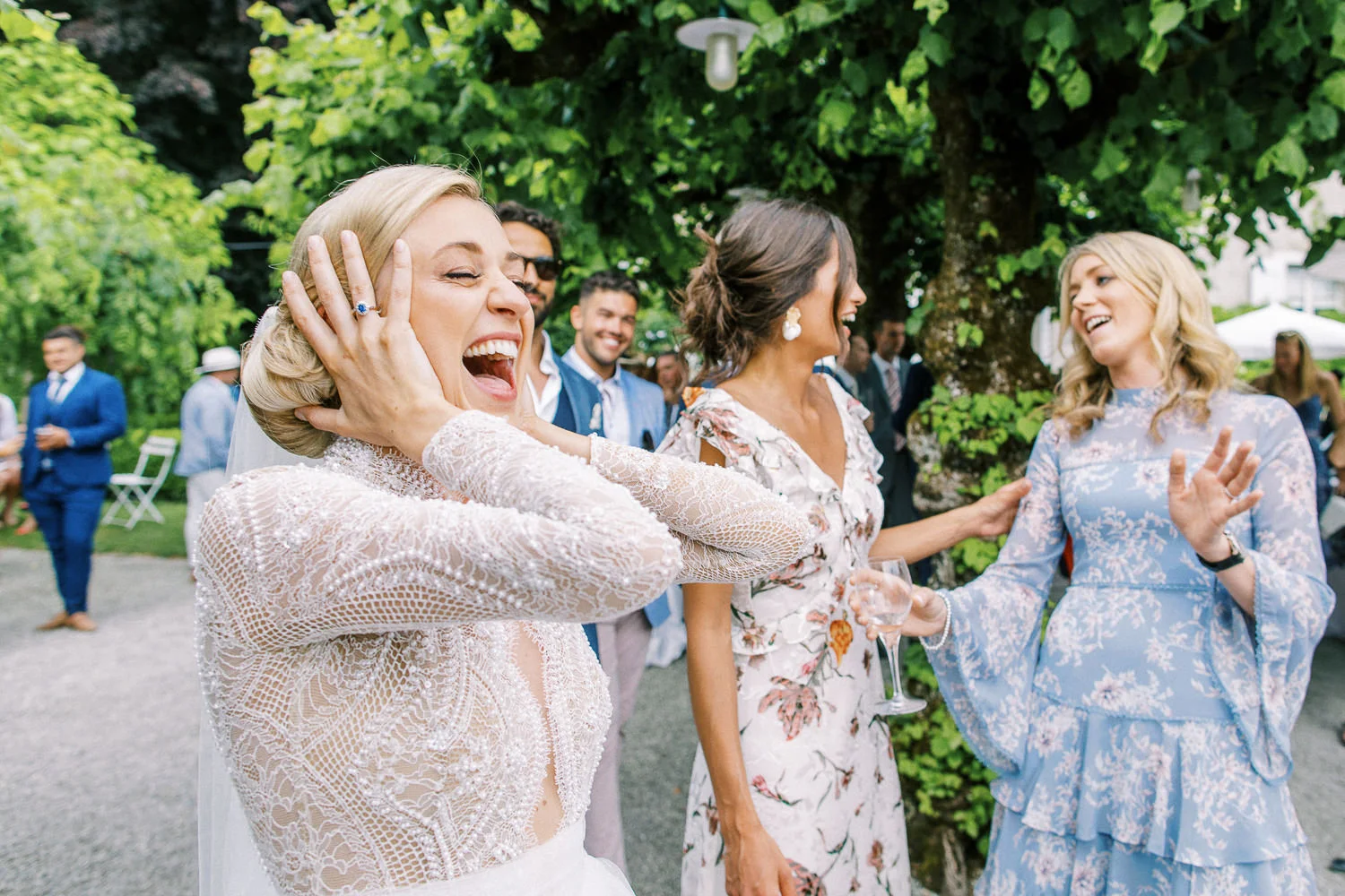 A bride in a lace gown laughs with pure joy, holding her hands to her head in surprise while celebrating with her friends.