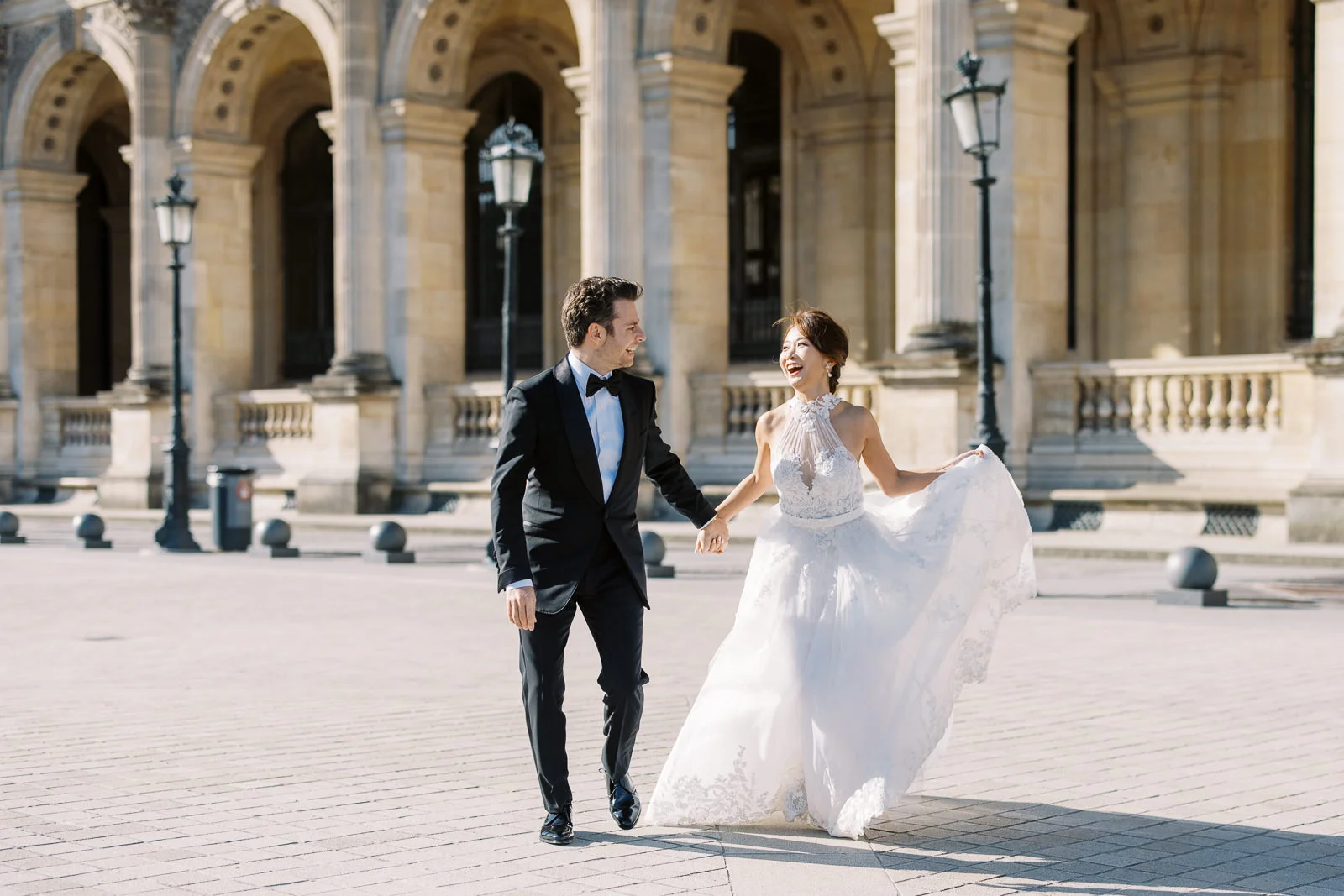 A bride and groom hold hands and smile at each other while walking through the courtyard of the Louvre Museum for their Paris couple photo session. How Much Does a Photographer in Paris Cost?