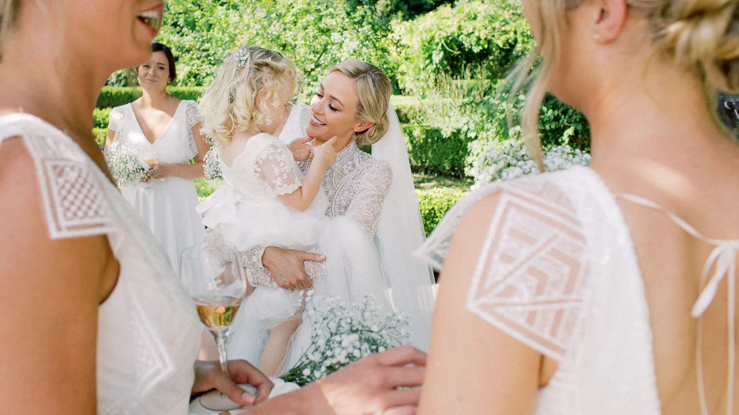 Joyful bride holding flower girl, surrounded by bridesmaids in a sunny garden, wedding photographer Lake Como. Photographe Mariage Lac de Côme.
