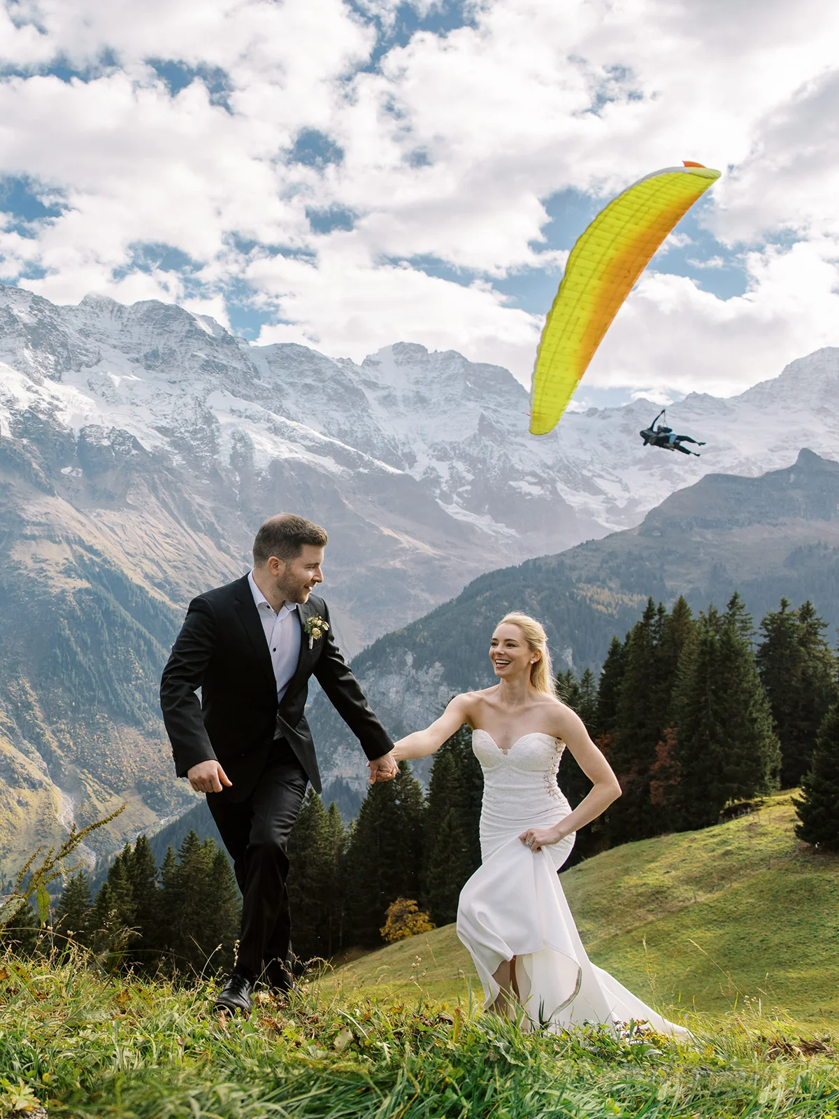 Bride and groom walking through Alpine meadows with a paraglider overhead by a Davos wedding photographer. Photographe mariage Davos.