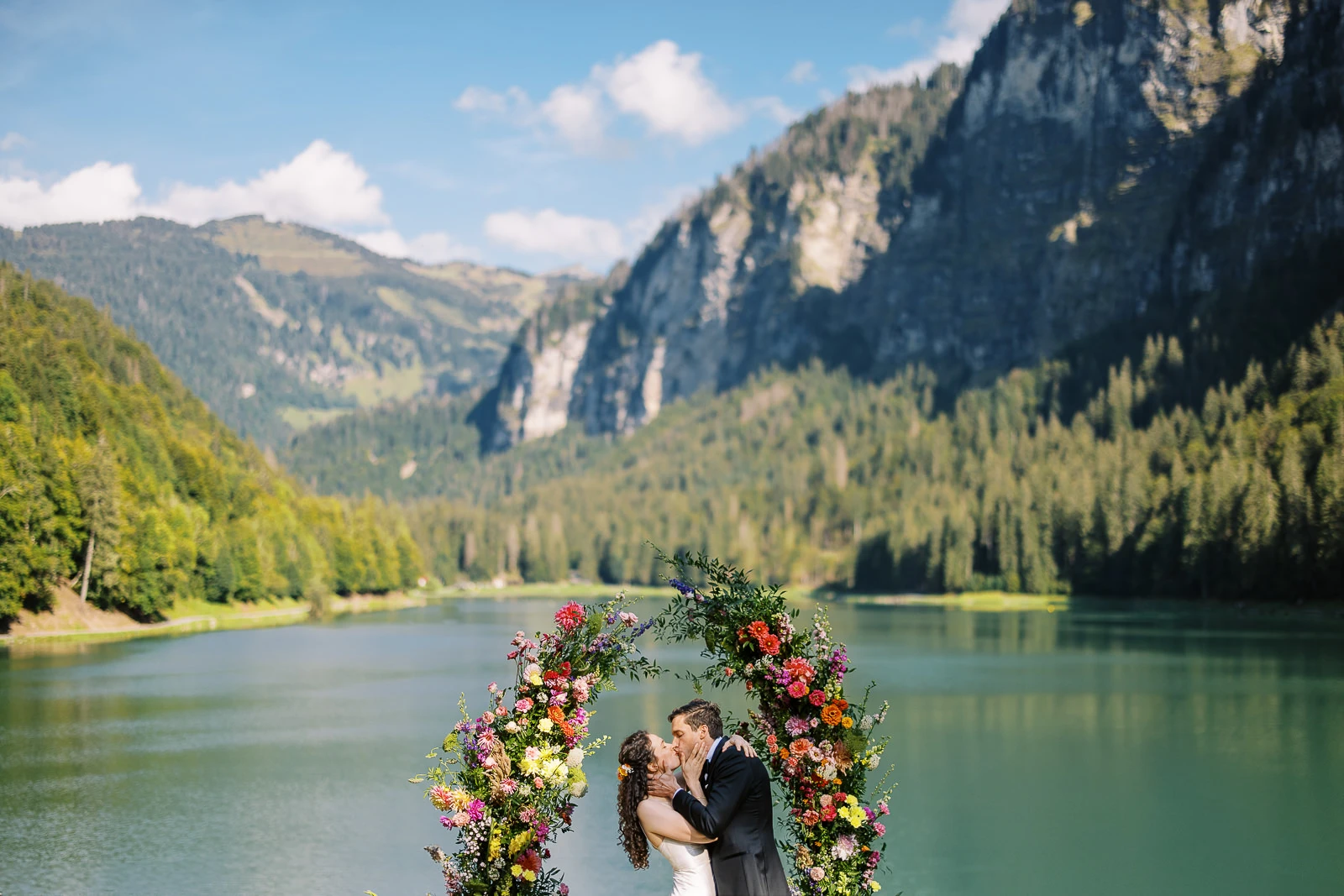 Couple kissing under floral arch by serene lake with majestic French Alps mountains, captured by a wedding photographer French Alps.