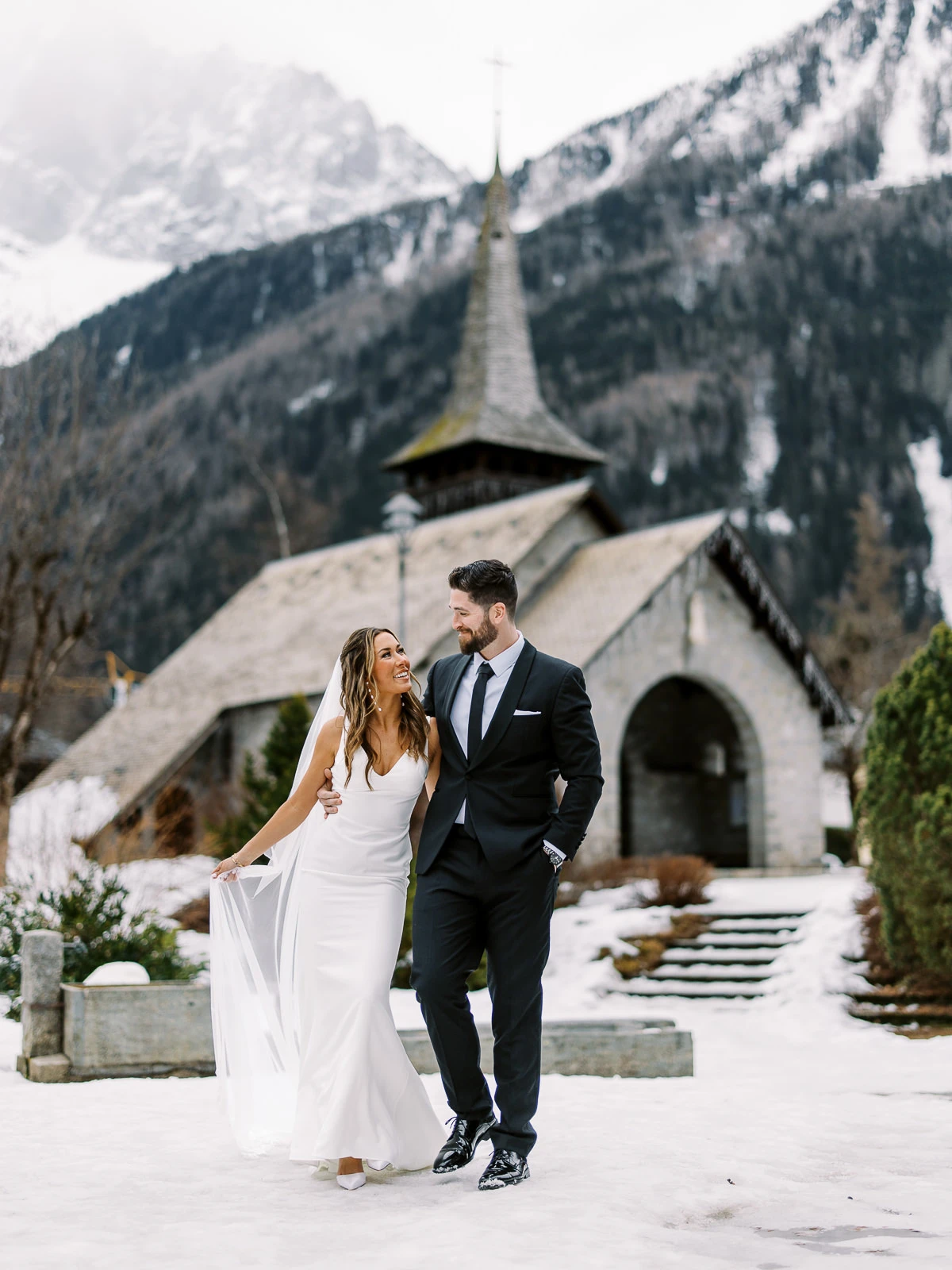 Couple walking in snow by a rustic church in Chamonix, captured by wedding photographer French Alps.