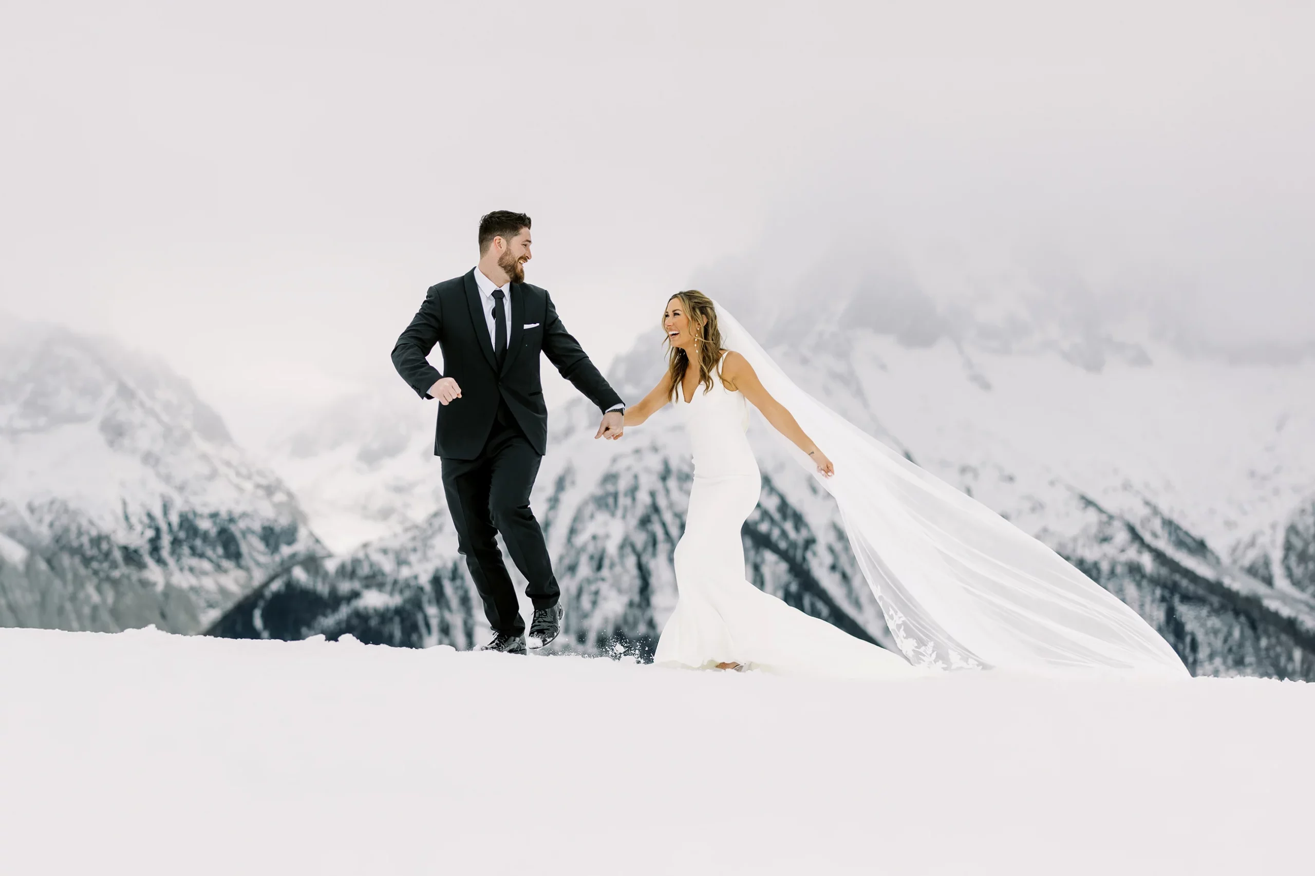 Joyful bride and groom running through fresh snow with majestic mountains, captured by wedding photographer Gstaad. Photographe mariage Gstaad.