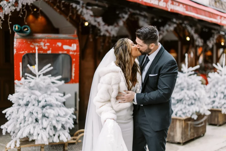 Couple wedding photographer capturing a romantic kiss amidst snowy trees and festive village decor.