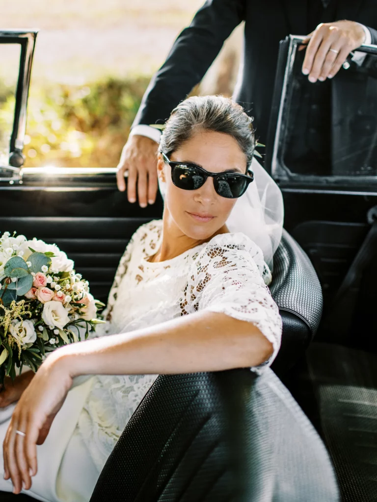 Bride wearing sunglasses in a vintage car by a french wedding photographer.