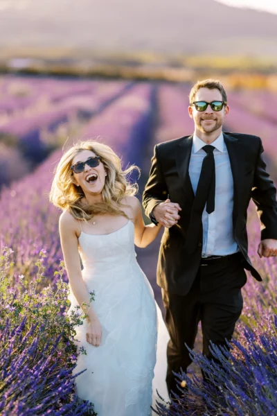 Bride and groom wearing sunglasses running through lavender fields by a French wedding photographer.