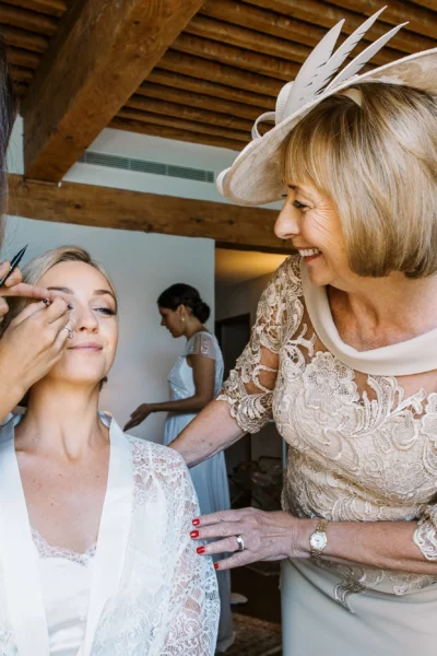 Bride receiving makeup finishing touches while her mother watches, captured by a French wedding photographer.