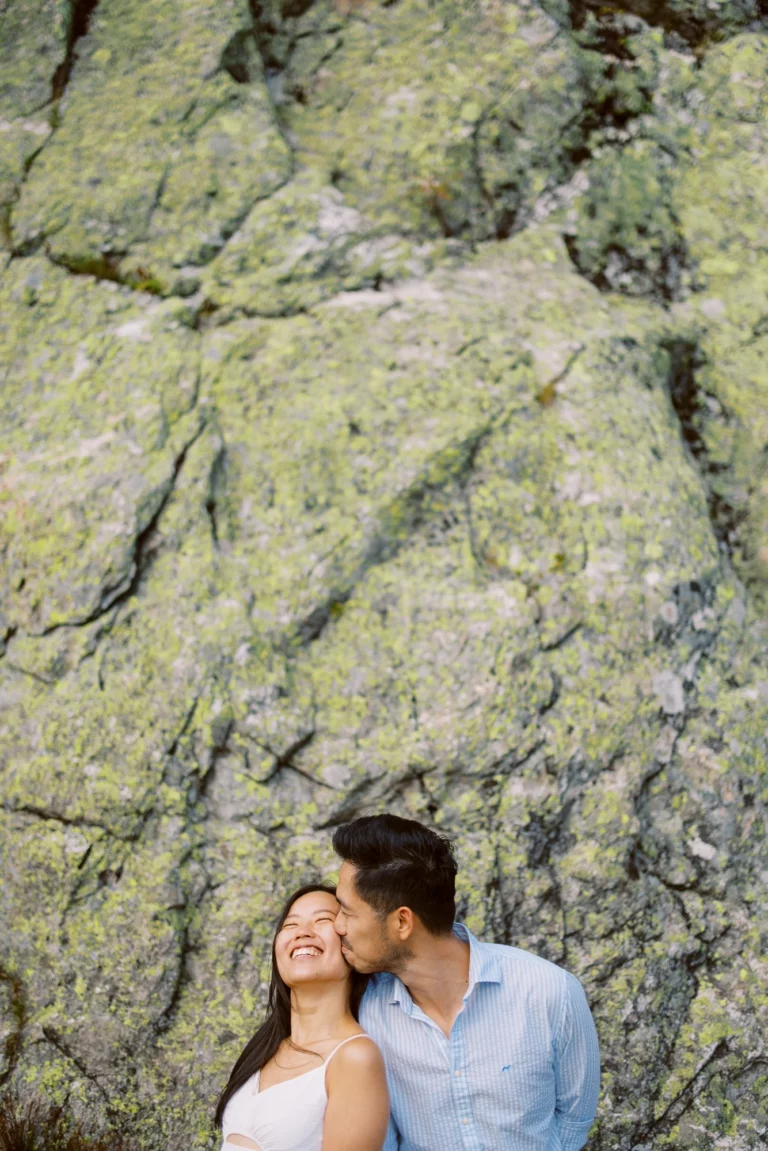 Couple joyeux riant lors d'une séance engagement devant des rochers moussus. Photographe de mariage.