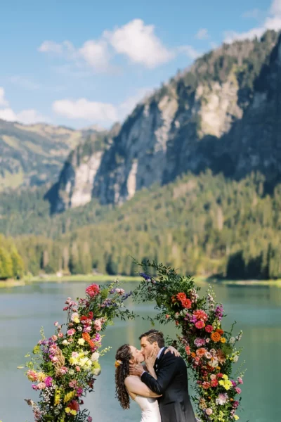 A romantic kiss under a floral arch at an alpine lake, by a French wedding photographer.