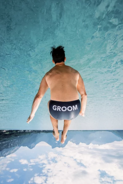 A groom jumping into crystal clear turquoise water in the Alps by a French wedding photographer.