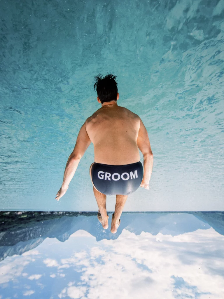 A groom jumping into crystal clear turquoise water in the Alps by a French wedding photographer.