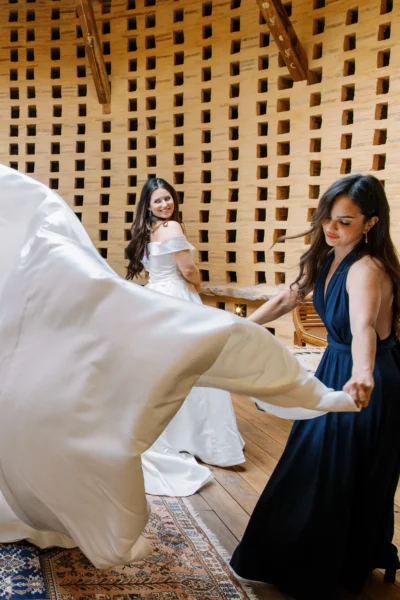 A bride smiles as her friend tosses her gown train, captured by a French wedding photographer.