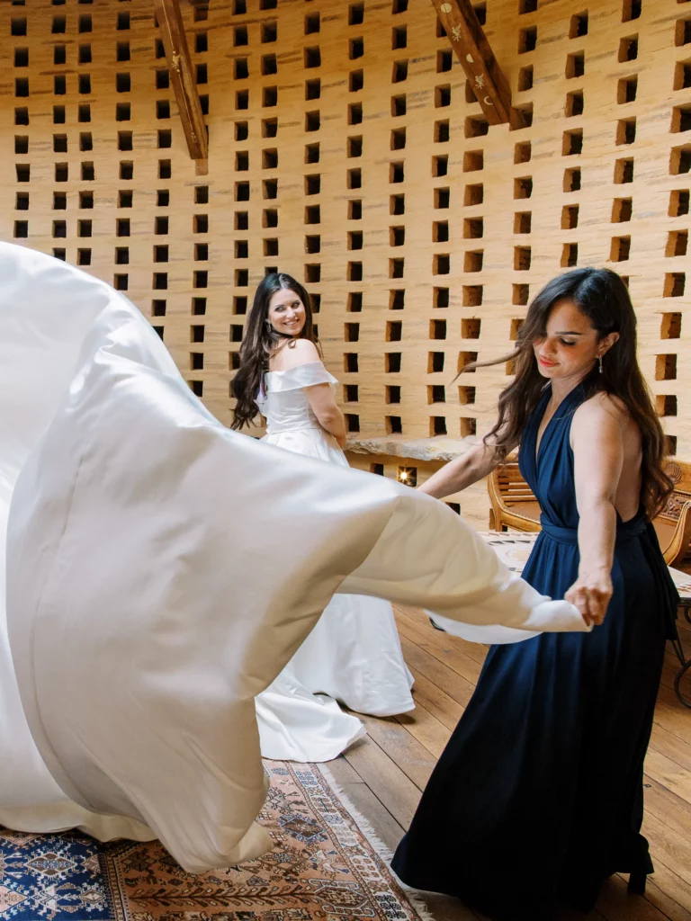A bride smiles as her friend tosses her gown train, captured by a French wedding photographer.